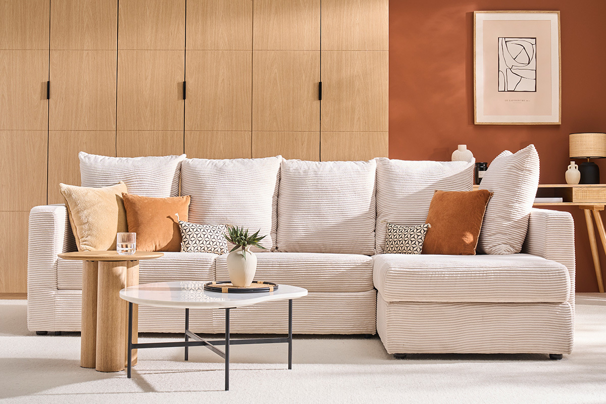 Divano letto ad angolo in velluto a coste beige, cuscini abbinati, tavolino e pianta, sfondo in legno chiaro.