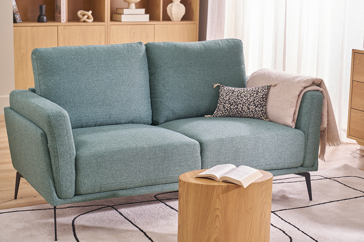 Divano a 2 posti verde grigio con piedi neri, vista da 3/4 in un soggiorno.