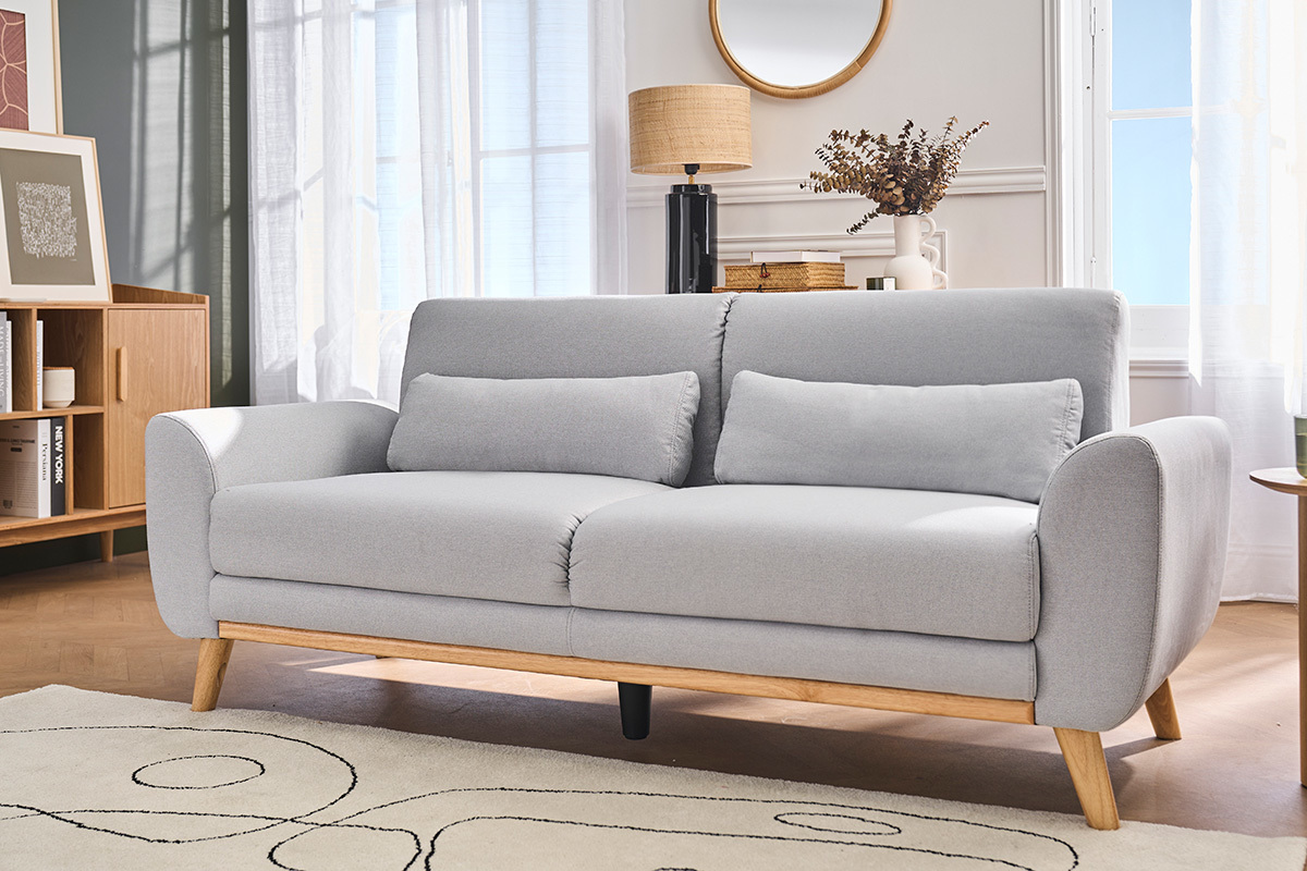 Divano scandinavo a 3 posti, tessuto grigio chiaro, piedi in legno chiaro, vista da 3/4 in un salotto.