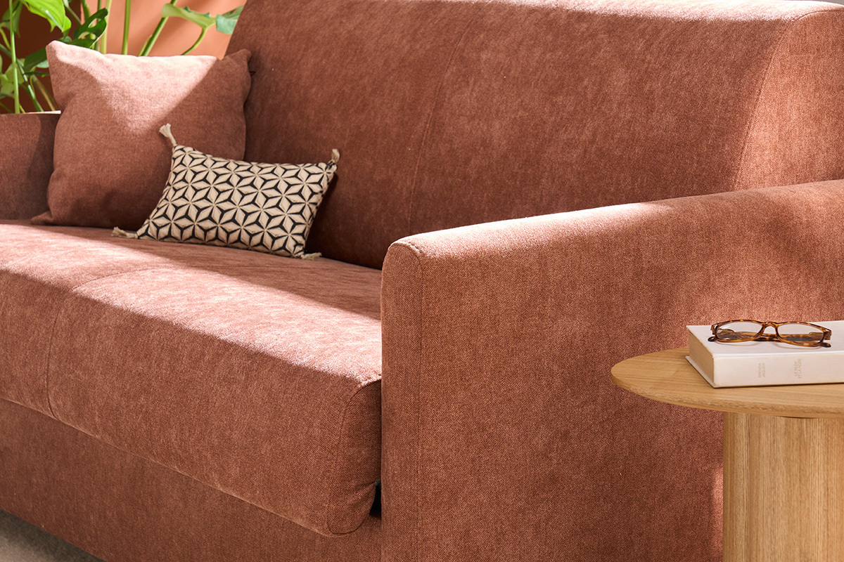 Divano letto in velluto terra bruciata, bracciolo destro e cuscini con tavolino in legno.