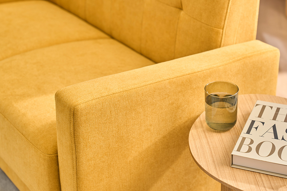 Divano a 2 posti in velluto di colore giallo senape, bracciolo in primo piano, tavolino con bicchiere e libro.