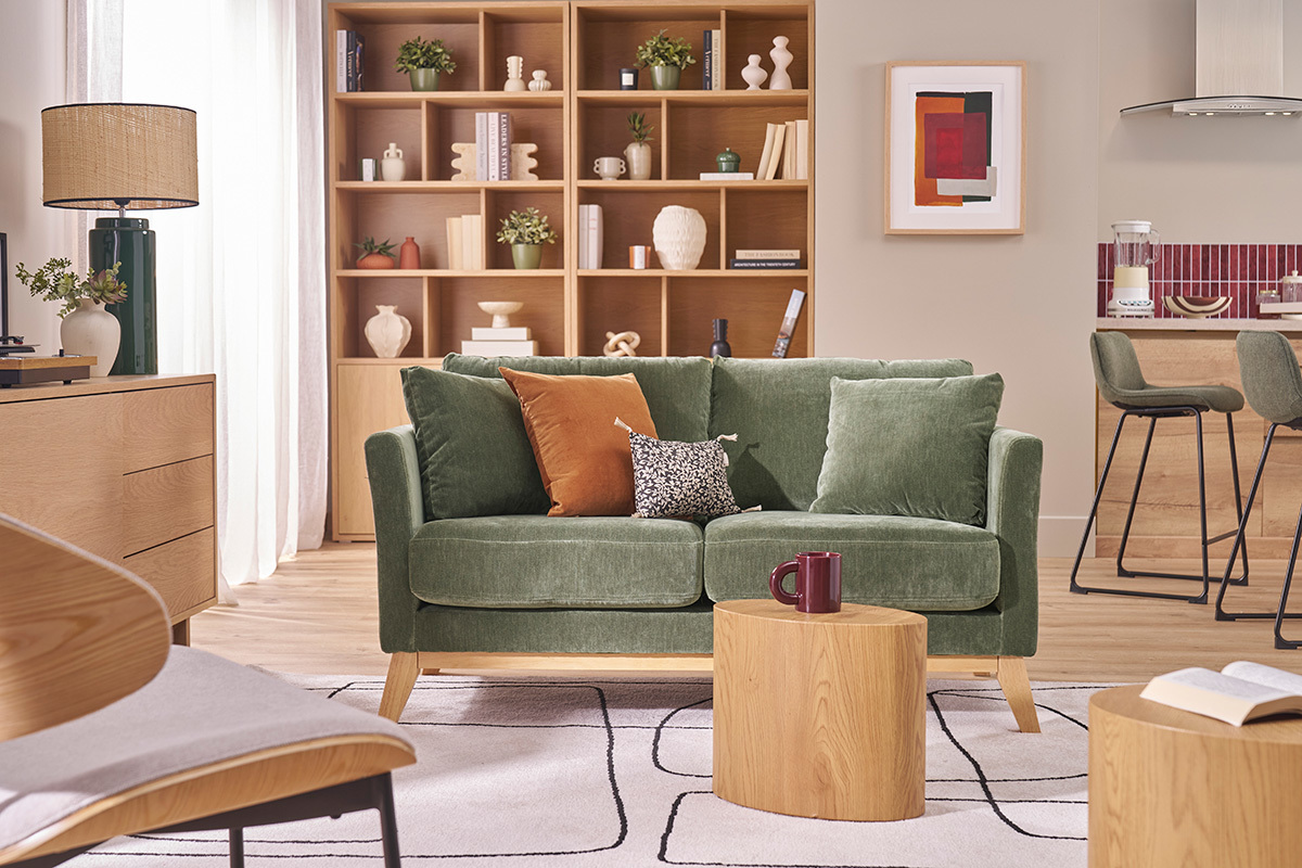 Divano scandinavo verde kaki Oslo a 2 posti, con piedi in legno, in un soggiorno con tavolo in legno e scaffali.