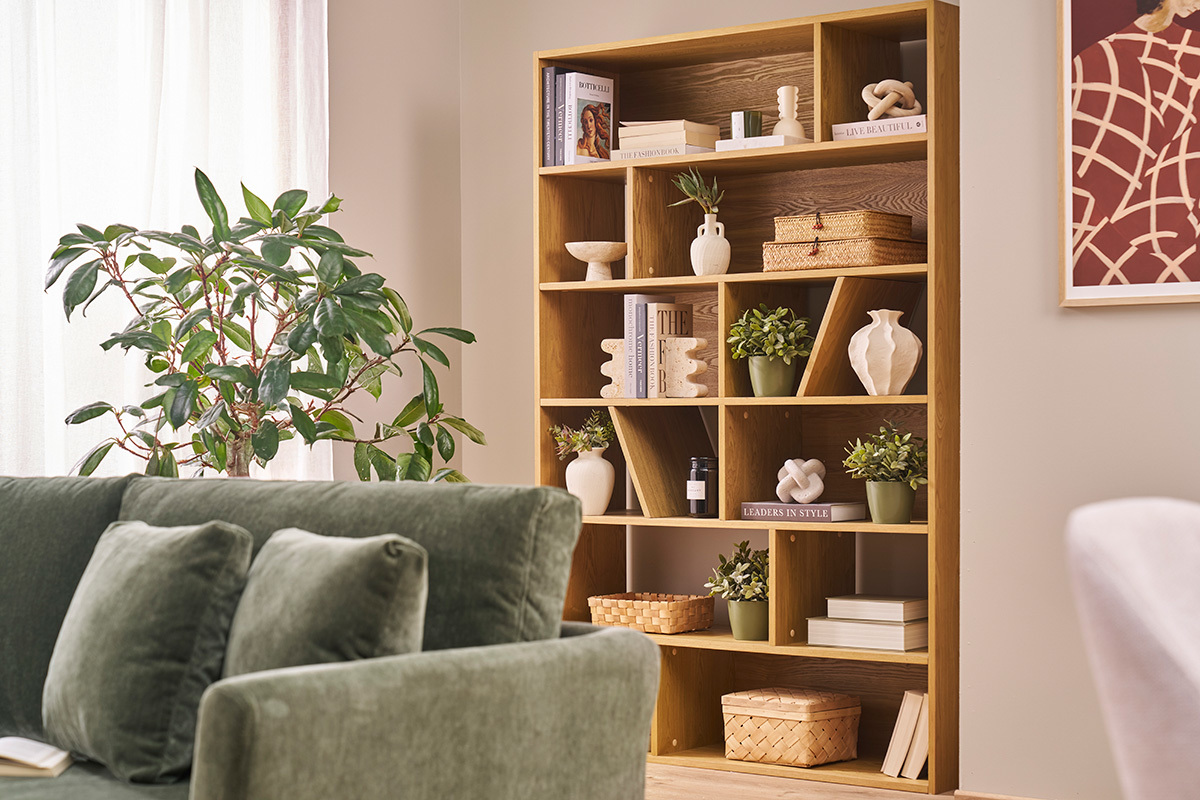 Libreria in legno chiaro con ripiani asimmetrici, decorata con libri e piante, vista di lato.
