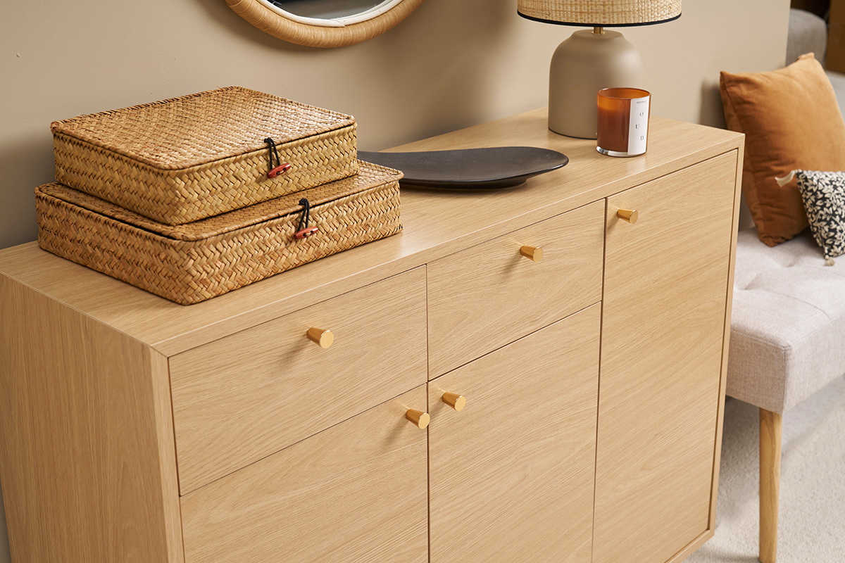 Mobile da ingresso in legno chiaro con maniglie dorate, due cesti in vimini e decorazioni sulla parte superiore.