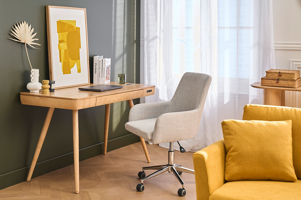Sedia da ufficio in tessuto grigio chiaro SHANA davanti a una scrivania in legno, accanto a un divano giallo.