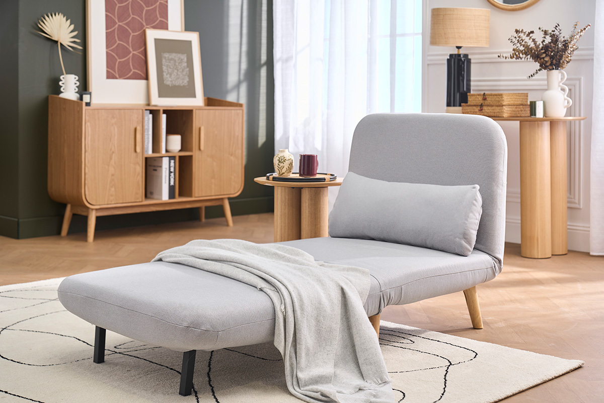 Poltrona trasformabile AMIKO in tessuto grigio chiaro, vista 3/4, con plaid, tavolino e decorazione scandinava.