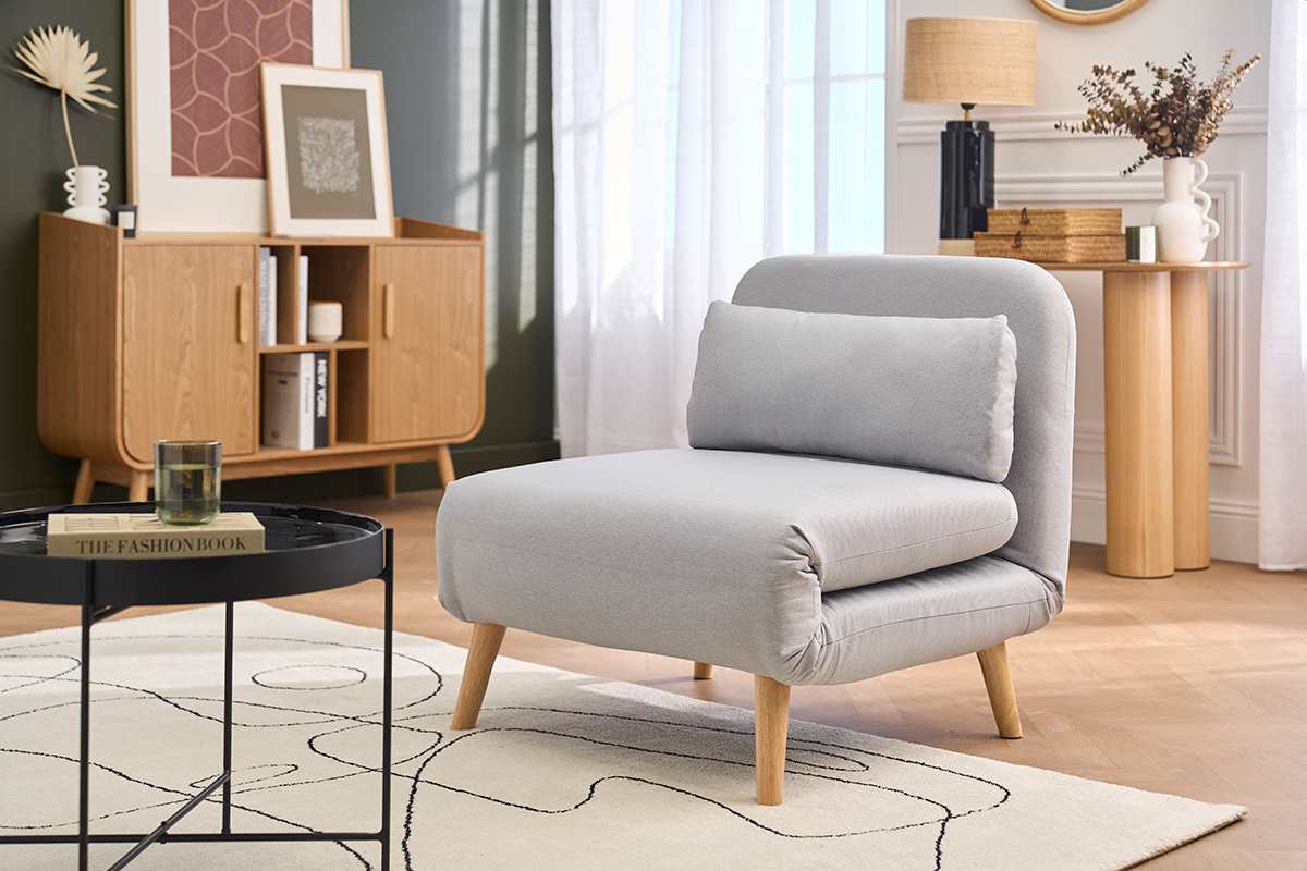 Poltrona convertibile in tessuto grigio chiaro e legno chiaro, vista da 3/4 in un salotto elegante.