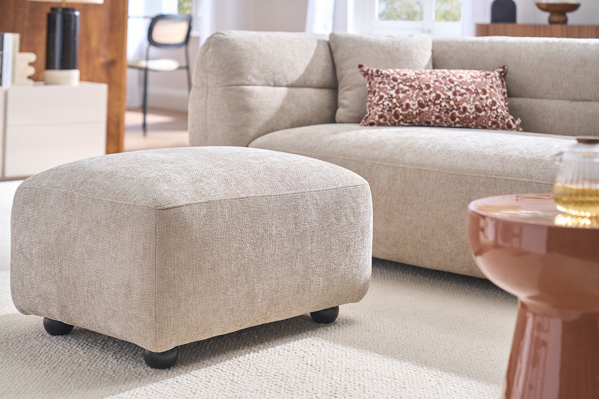 Pouf poggiapiedi in tessuto beige, con divano sullo sfondo, vista a 3/4