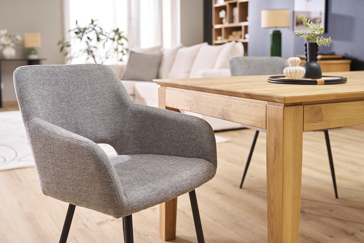 Sedia in tessuto effetto velluto grigio con gambe in metallo nero, vista da 3/4 vicino a un tavolo in legno.