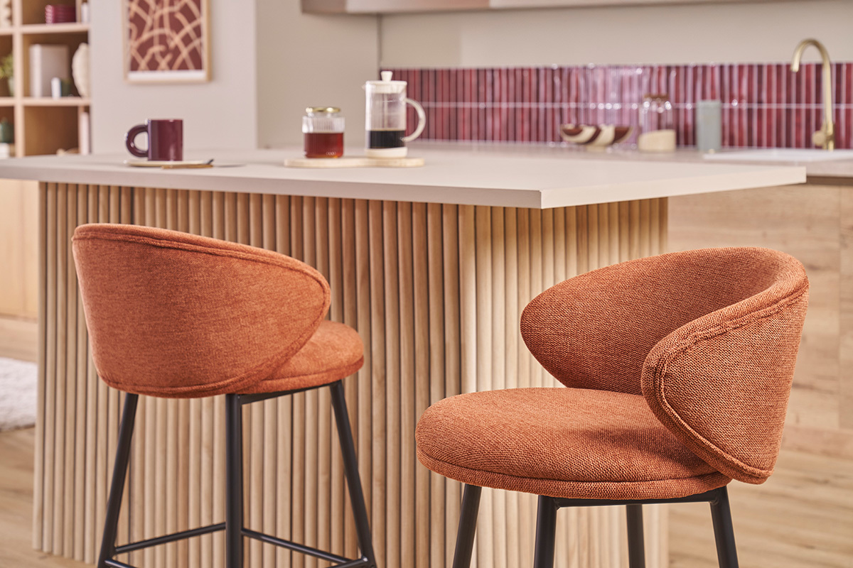 Due sgabelli da bar in tessuto color terra bruciata con piedi metallici neri davanti a un'isola da cucina.