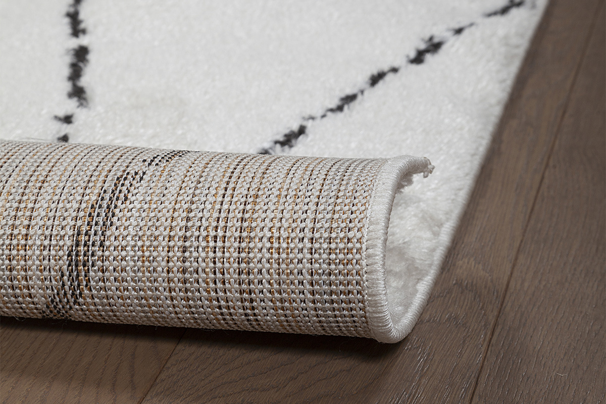 Primo piano sul tappeto SILAN con motivi neri e texture in tessuto, parzialmente arrotolato.