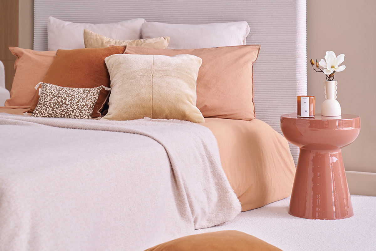 Tavolino rotondo in metallo smaltato terracotta accanto a un letto con cuscini beige e terracotta.