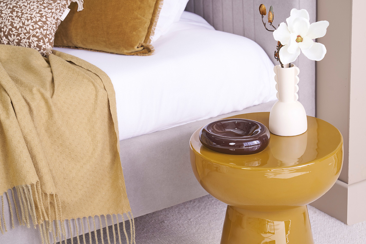 Tavolino da appoggio MOGOU rotondo bronzo con vaso bianco e portacenere marrone, accanto a un letto.