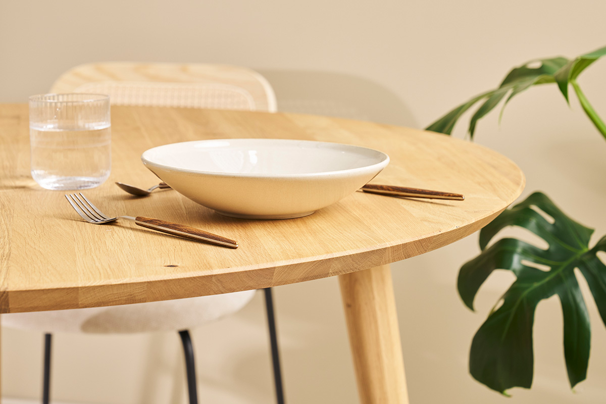 Tavolo rotondo in rovere chiaro con stoviglie e pianta sullo sfondo, vista ravvicinata.